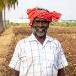 Portrait of a smiling South Asian farmer with a red headscarf, standing in a sunlit field.