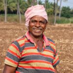 A joyful senior farmer wearing a pink turban stands in a ploughed field under palm trees.